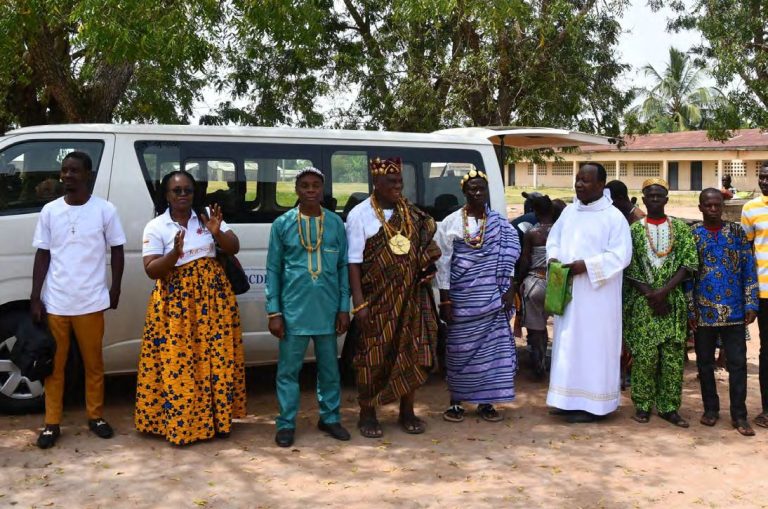 Une délégation de Caritas, dont un prêtre en aube et des chefs traditionnels en tenue de cérémonie, devant un minibus.