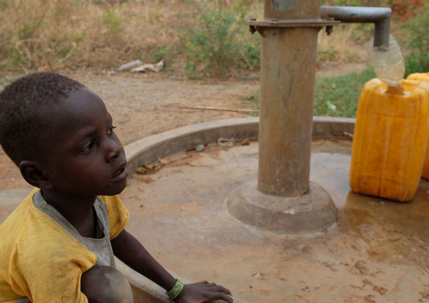 Un enfant attend près d'une pompe à eau où se remplit un bidon jaune.