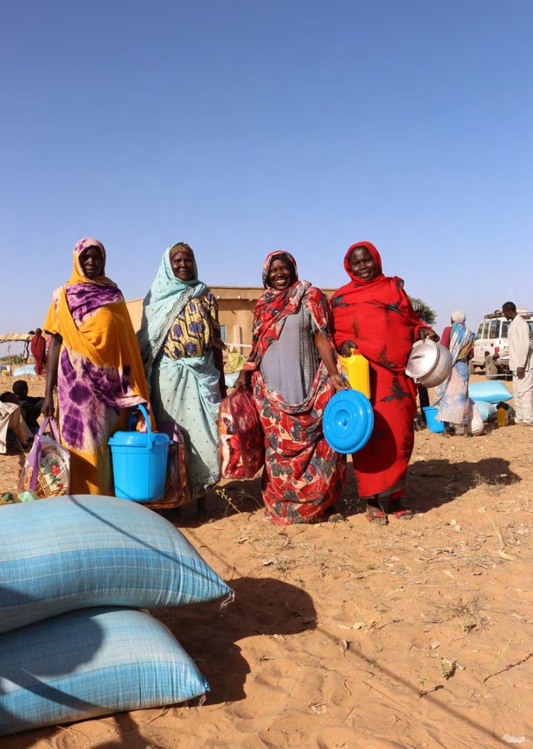 Des femmes en voiles colorés reçoivent des seaux et des nattes lors d'une distribution, dans une zone sablonneuse.
